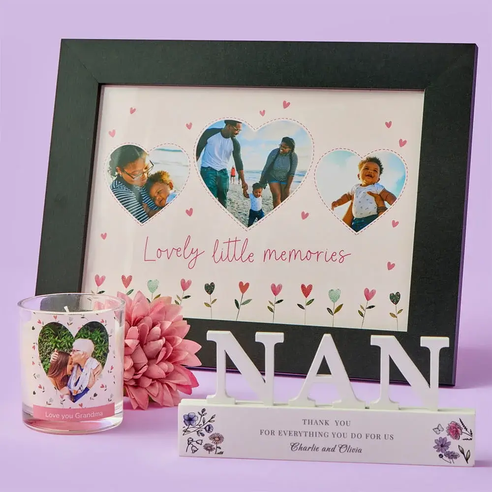 A heart‑themed photo frame with three family photos sits behind a personalised candle and a decorative NAN ornament, all arranged with a pink flower on a pastel purple background.